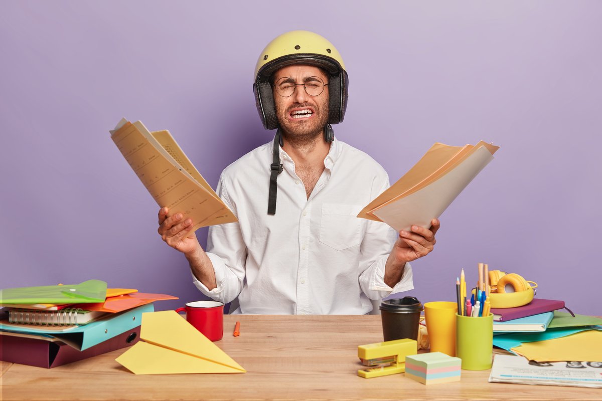 Frustrated overworked male office worker holds papers in both hands, cries from despair, wears spectacles, protective headgear, poses at desktop with stationary around, isolated on purple wall